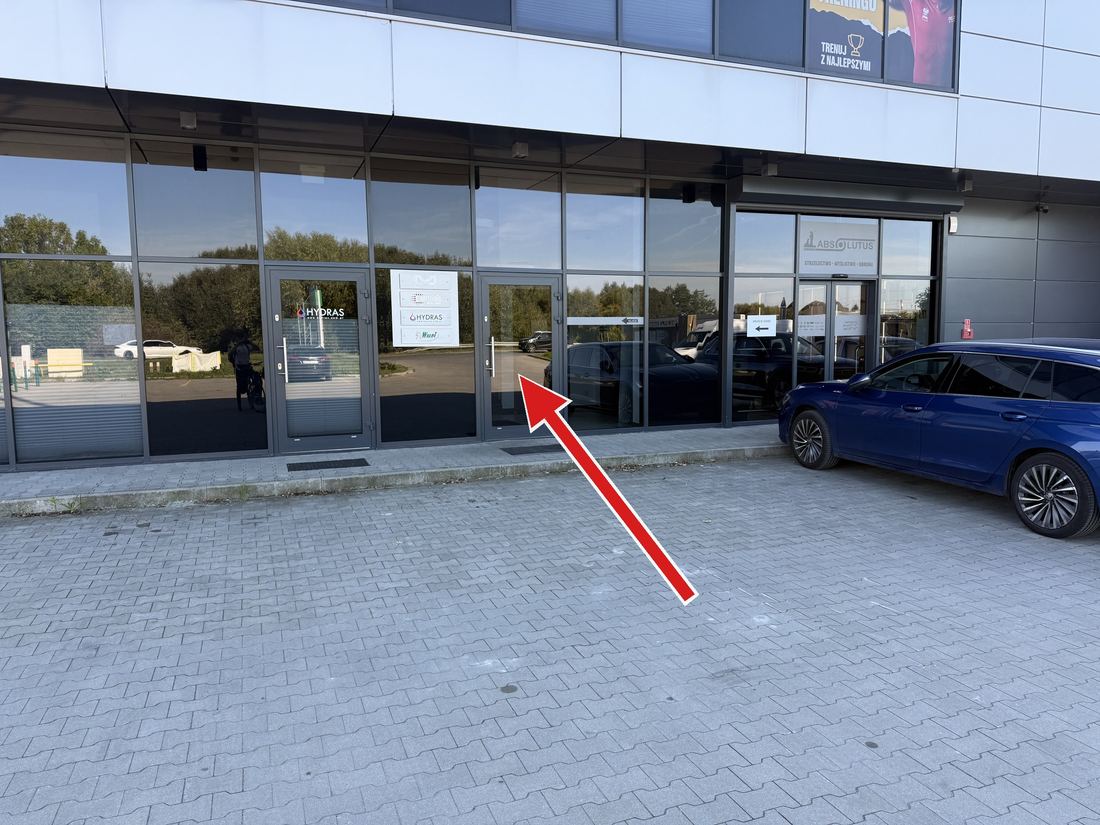 Wejście dla klientów Dom Siły — drzwi na prawo od białej tablicy z szyldami, widok z parkingu